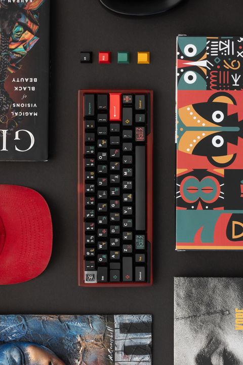 Black mechanical keyboard on a dark desk surrounded by art books, keycaps, and a red cap
