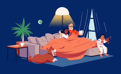 A family under a big orange blanket on the sofa on a winter evening, a child with a mug beside them, a dog on the rug, a lamp glowing and snow outside the window