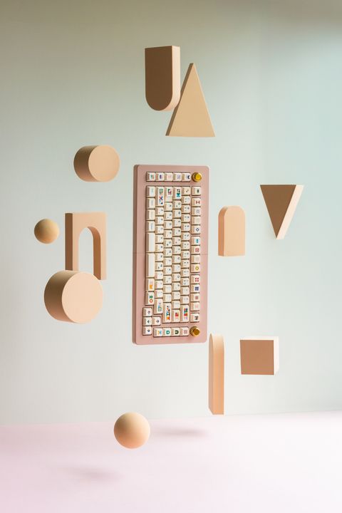 A pink split keyboard fitted with Abstract, its two halves joined and floating among peach geometric shapes: arches, cylinders, triangles, a cube and spheres