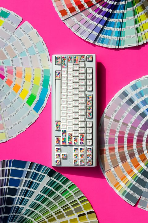 White mechanical keyboard with a colorful abstract keycap set on a hot pink background surrounded by fanned Pantone swatch books